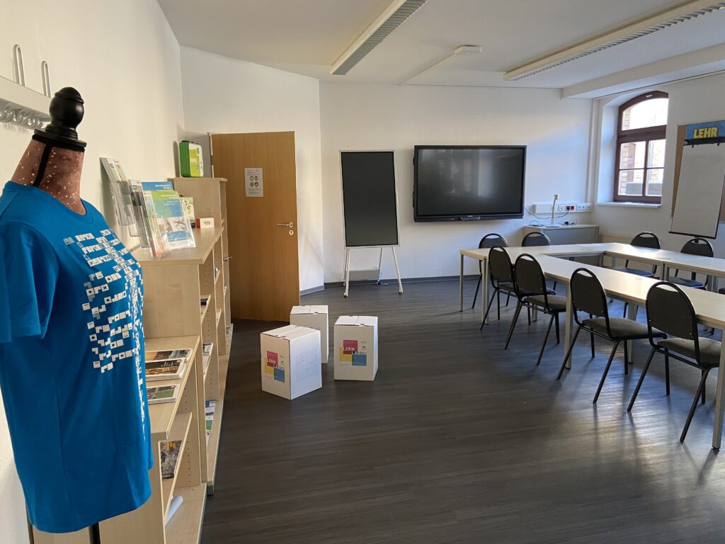 Hier ein Blick in das Digitale Lernlabor: Es ist ein hellen Raum, zu sehen sind Regale mit Informationsmaterialien, Seminar-Tische und Stühle, eine mobile Flipchart und an der Wand befindet sich eine interaktive Tafel.